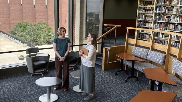 two women standing among tables, chairs, and benches with shelves of library books in the background