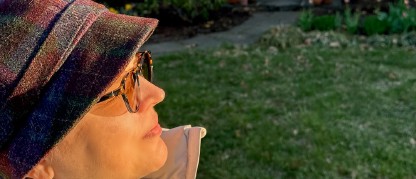 Woman in a winter hat and coat outdoors in front of a house facing the rising sun.
