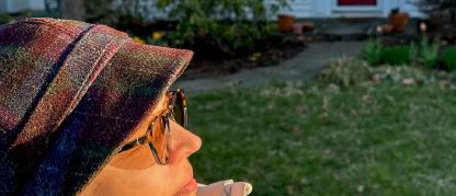 Woman in a winter hat and coat outdoors in front of a house facing the rising sun.